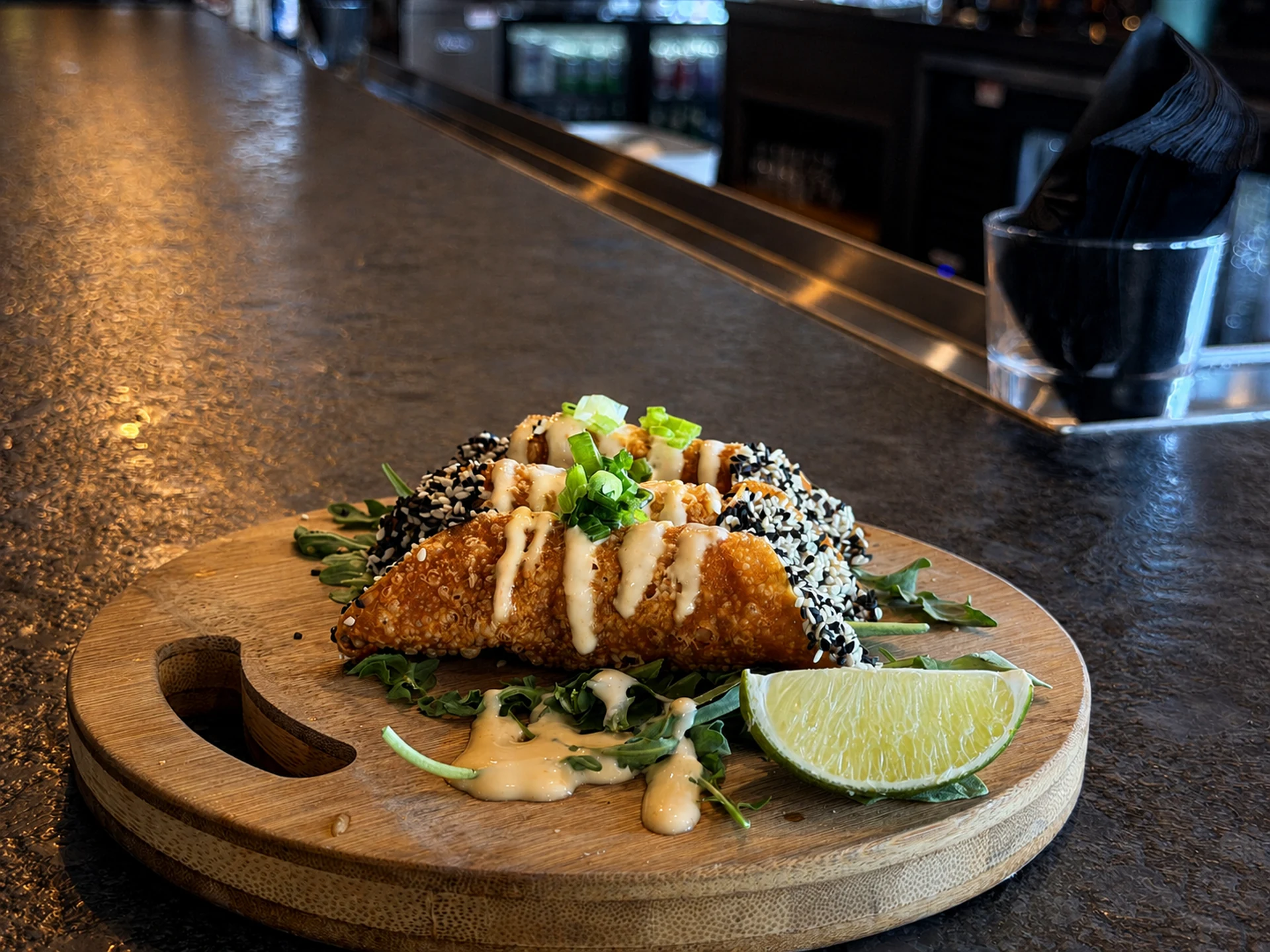 Crisp sesame crusted shells on a round wood board with scallion, drizzled aioli and a lime wedge on the bar top