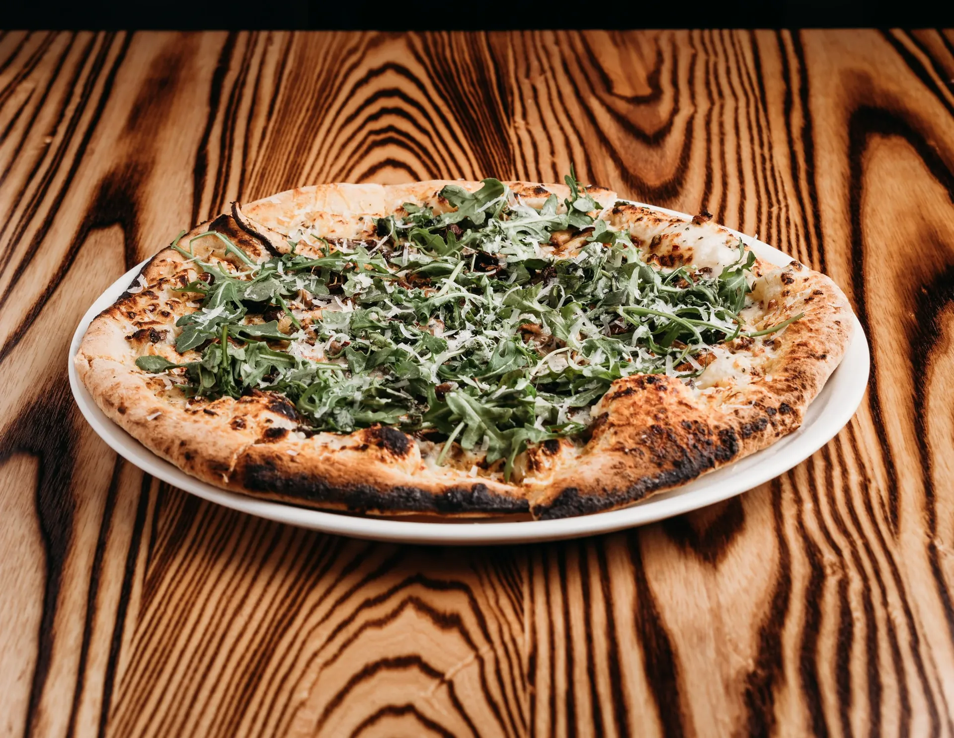 Wood-fired pizza topped with arugula and shaved parmesan on a wood table